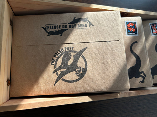 Brown envelopes with 'The Paleo Post' logo in a wooden box.
